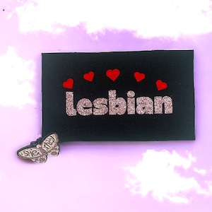Puede incluir: Un parche negro con la palabra "lesbiana" en letras de purpurina rosa y cinco corazones rojos sobre la palabra. El parche está sobre un fondo rosa con nubes blancas.