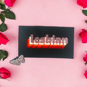 Puede incluir: Una tarjeta negra con la palabra "lesbiana" en letras de purpurina rosa sobre un fondo rosa rodeado de rosas rojas.