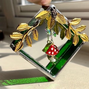 May include: A green glass square with gold leaf branches and a red and white mushroom hanging from it. The mushroom is attached to a chain with clear and iridescent beads.