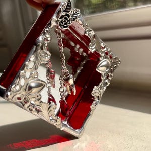 May include: A small, red glass box with silver filigree detailing. The box has a hinged lid and is decorated with silver charms, including a tooth and a rose.