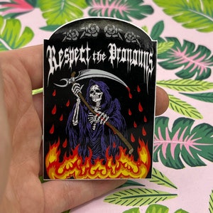 May include: A sticker featuring a Grim Reaper holding a scythe, standing in flames with red drops of blood falling from the sky. The text "Respect the Pronouns" is written in a gothic font at the top of the sticker.