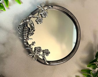 May include: A silver hand mirror with a floral design and a scorpion on the side. The mirror is oval shaped and has a silver frame.