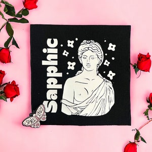 Könnte beinhalten: Ein schwarzer Stoff-Patch mit weißem Text, der "Sapphic" lautet, und einer Strichzeichnung einer Frau mit Blumen. Der Patch befindet sich auf einem rosa Hintergrund mit roten Rosen.
