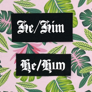 May include: Two black fabric patches with white text that reads "He/Him" in a gothic font. The patches are on a pink background with a tropical leaf pattern.