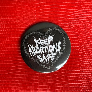 Puede incluir: Un botón negro y blanco con un diseño de corazón de alambre de púas. El texto dentro del corazón dice "Keep Abortions Safe".