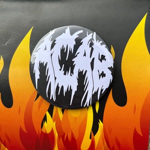 May include: A black and white button with the word "ACAB" in a stylised font. The button is on a background of orange and yellow flames.