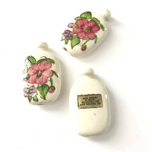 May include: Three off-white ceramic perfume bottles. Two bottles are decorated with pink and purple floral designs and green leaves. The third bottle has a label that reads "Hand Painted in Brazil exclusively for Avon Products, Inc."