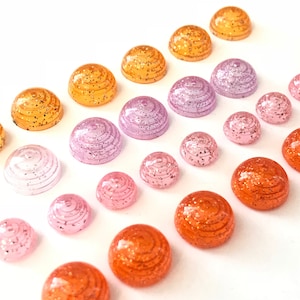May include: An assortment of colorful, dome-shaped embellishments in shades of orange, pink, and purple. Each has a spiral design and glitter accents. These craft supplies are arranged on a white background.