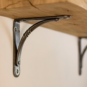 Può includere: Primo piano di una mensola in legno rustico sostenuta da una staffa decorativa in metallo scuro. La staffa ha un design curvo con un dettaglio attorcigliato ed è fissata alla mensola con viti. La mensola è in legno chiaro con venature visibili.