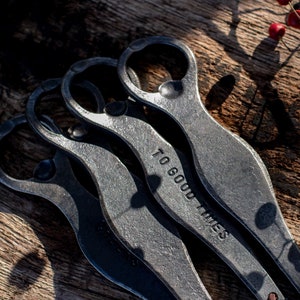 Puede incluir: Tres abridores de botellas de plata apilados uno encima del otro sobre una superficie de madera. El abridor superior tiene las palabras "To Good Times" grabadas.