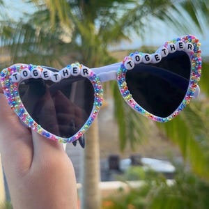 May include: Heart-shaped sunglasses with white frames featuring iridescent, multi-colored rhinestones along the rims. The top edges of the frames are decorated with white letter beads spelling out the word TOGETHER across both lenses.
