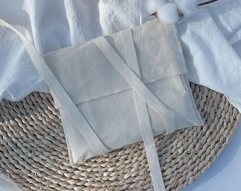May include: A neutral-toned, rectangular pouch with a flap closure and a woven strap. The pouch is made of a soft, textured fabric. The strap is a light beige color with a subtle herringbone pattern. The pouch is resting on a woven mat.