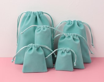 Pode incluir: Um conjunto de seis sacos de veludo azul-petróleo com cordão, de tamanhos variados. Cada saco tem um cordão a condizer. Fundo rosa e branco.