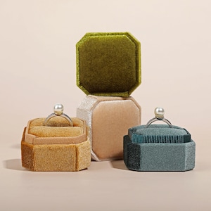 Puede incluir: Tres cajas de anillos de terciopelo en diferentes colores. Las cajas están apiladas una encima de la otra. La caja superior es verde, la caja del medio es beige y la caja inferior es amarillo mostaza. Cada caja tiene un anillo dentro.