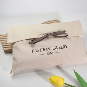 Pode incluir: Uma bolsa de joias de tecido bege com o texto "FASHION JEWELRY - JUNE" impresso em preto. A bolsa é amarrada com uma fita marrom e está parcialmente aberta. Uma tulipa amarela e uma peça decorativa de madeira estão no fundo.