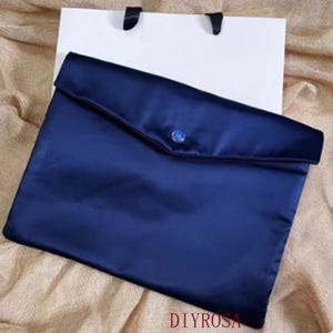 Op de afbeelding: Een marineblauwe satijnen clutch met een flap en een decoratieve knoop. Het staat tegen een witte boodschappentas met zwarte handvatten. De tekst "DIYROSA" is rechtsonder zichtbaar.