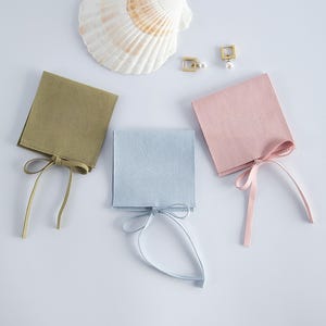 Pode incluir: Três pequenos sacos quadrados para joias em verde oliva, azul claro e rosa, cada um com uma fita a condizer. Brincos quadrados dourados com detalhes de pérolas e uma concha também são visíveis.