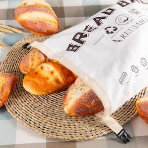 Puede incluir: Una bolsa de lona blanca con un logotipo marrón de "BREAD" y un cierre de cremallera. La bolsa está llena de diferentes panes, incluyendo una hogaza redonda, una baguette y una hogaza más pequeña. La bolsa está sentada sobre una estera de mimbre tejida.