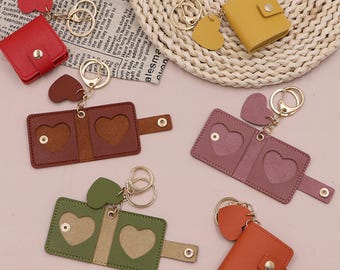 May include: A collection of leather keychains in various colors, including red, yellow, brown, purple, green, and orange. Each keychain features a heart-shaped charm and a small wallet or photo holder with heart-shaped cutouts. The keychains have gold-colored metal rings.