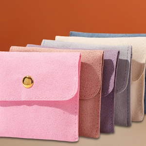 Puede incluir: Una colección de seis pequeñas bolsas de material similar al ante, cada una con una solapa y un botón dorado. Las bolsas son de varios colores pastel, incluyendo rosa, marrón, morado, gris, beige y azul.