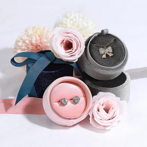 May include: Three velvet jewellery boxes with lids. The boxes are in shades of pink, blue, and grey. The pink box contains a pair of earrings, the blue box has a ribbon, and the grey box contains a necklace with a bow pendant.