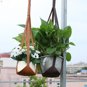 May include: Two hanging planters with leather straps. One holds white flowers, the other green plants. The planters are suspended from a metal pole. The plants are in pots.