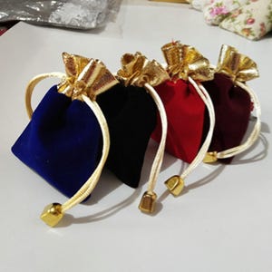 Puede incluir: Cuatro pequeñas bolsas de terciopelo con cordón de ajuste en azul, negro, rojo y burdeos con ribetes dorados. Las bolsas están atadas con cordones blancos y tienen borlas doradas.