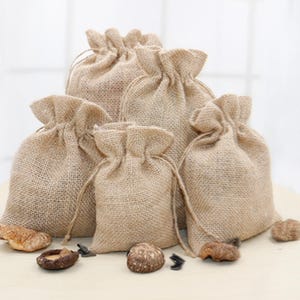 Könnte beinhalten: Satz mit fünf kleinen, natürlichen Jutesäcken mit Kordelzug. Die Säcke bestehen aus einem groben, gewebten Stoff und haben ein rustikales, erdiges Aussehen. Sie sind perfekt zum Verpacken von kleinen Geschenken oder Mitbringseln.