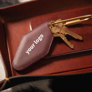 May include: A burgundy leather keychain with the text "your logo" in white, attached to a brass key ring and two gold keys. The keychain rests on a dark red leather tray.