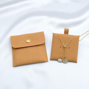 May include: Two tan jewelry pouches, one closed with a gold snap and the other open, displaying a gold necklace with a fan-shaped pendant. The necklace has a delicate chain and a white and crystal pendant.
