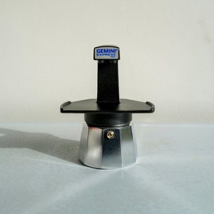 Op de afbeelding: Een zilveren en zwarte Gemini Express espressomachine met een zwarte handgreep en een zilveren basis. De machine heeft een blauw label met de tekst "GEMINI EXPRESS ITALY".