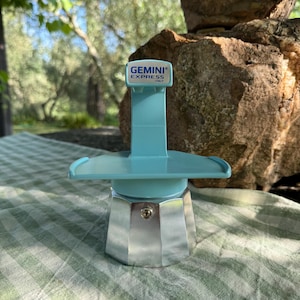 May include: A silver and blue Gemini Express coffee maker with a blue plastic stand. The stand has a label that reads "GEMINI EXPRESS ITALY".