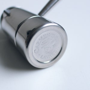 May include: A polished stainless steel stovetop espresso maker with a cylindrical body and a handle. The base features an engraved logo that reads Carmencita Lavazza, along with technical markings including Mod. 24A and Made in Italy.