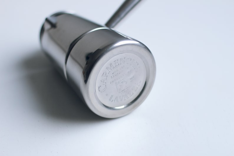 May include: A polished stainless steel stovetop espresso maker with a cylindrical body and a handle. The base features an engraved logo that reads Carmencita Lavazza, along with technical markings including Mod. 24A and Made in Italy.