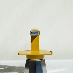 Op de afbeelding: Een gele en zilveren espressomaker met de tekst "GEMINI EXPRESS ITALY" erop. De koffiemaker heeft een zeshoekige basis en een gele handgreep en deksel. De afbeelding is gemaakt in natuurlijk licht.