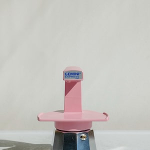 May include: A pink and silver Gemini Express stovetop espresso maker. The pink components include the top and handle, with the text "GEMINI EXPRESS ITALY" on the top. The base is silver. The espresso maker is designed for stovetop use.