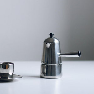 May include: A stainless steel stovetop espresso maker with a cylindrical body, a side handle, and a small black knob on the lid. Beside it sits a matching stainless steel espresso cup on a small saucer, both featuring a polished metallic finish.