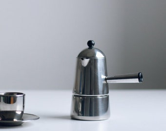 May include: A stainless steel stovetop espresso maker with a cylindrical body, a side handle, and a small black knob on the lid. Beside it sits a matching stainless steel espresso cup on a small saucer, both featuring a polished metallic finish.