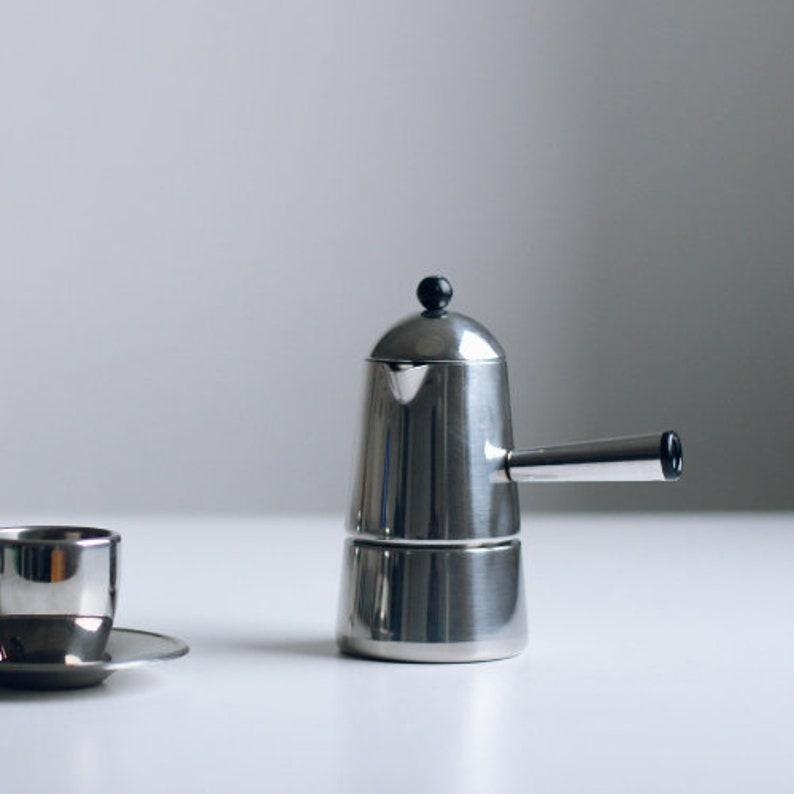 May include: A stainless steel stovetop espresso maker with a cylindrical body, a side handle, and a small black knob on the lid. Beside it sits a matching stainless steel espresso cup on a small saucer, both featuring a polished metallic finish.