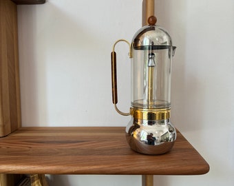 May include: A silver and gold coffee maker with a glass carafe sits on a wooden shelf. The coffee maker has a wooden handle.