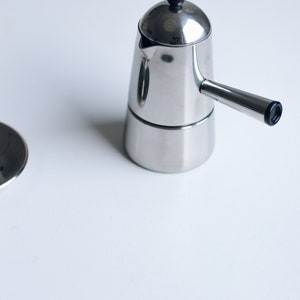 May include: A polished stainless steel stovetop espresso maker with a conical body, a side handle, and a small black knob on the lid. The coffee maker features a metallic finish and a simple design, accompanied by a separate circular metal lid component.