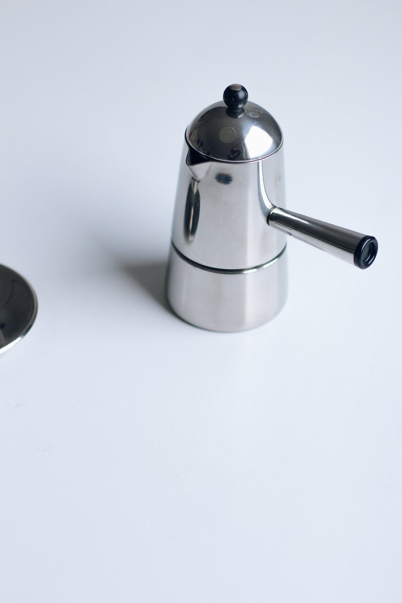 May include: A polished stainless steel stovetop espresso maker with a conical body, a side handle, and a small black knob on the lid. The coffee maker features a metallic finish and a simple design, accompanied by a separate circular metal lid component.