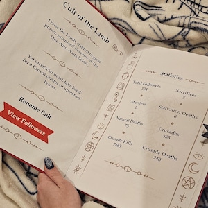 May include: A book with a red cover and the title "Cult of the Lamb". The book has a page open to a section titled "Statistics" with details about the cult, including the number of followers, sacrifices, murders, natural deaths, starvation deaths, crusades, crusade kills, and crusade deaths.