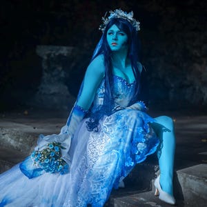 Könnte beinhalten: Eine Person in einem blau-weißen Hochzeitskleid mit blauer Haut und Haaren sitzt auf Steinstufen. Das Kleid hat Spitze und Blumendetails, und sie trägt einen Blumenkranz und einen Schleier. Sie hält einen blauen Strauß und trägt weiße Absätze.