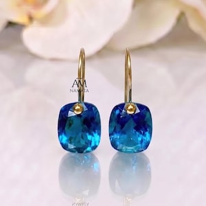 May include: A pair of gold-colored drop earrings featuring square, faceted, blue gemstones. The earrings have a simple design with a small gold ball connecting the gemstone to the gold hook. The earrings are displayed on a reflective surface.