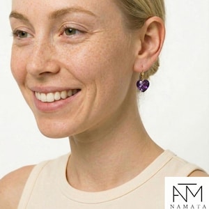 May include: A close-up of a person wearing a gold earring with a heart-shaped, deep purple gemstone. The earring is dangling from the ear. The person is smiling and wearing a cream-colored top.