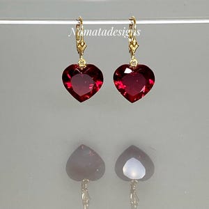 Puede incluir: Un par de pendientes en forma de corazón con una piedra preciosa de color rojo intenso. Los pendientes tienen un cierre de palanca de color dorado. Los pendientes se exhiben sobre una superficie reflectante.