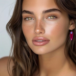 Puede incluir: Una mujer con el pelo largo y castaño y pecas lleva un par de pendientes colgantes. Los pendientes están hechos de un material rosa con un acento turquesa.