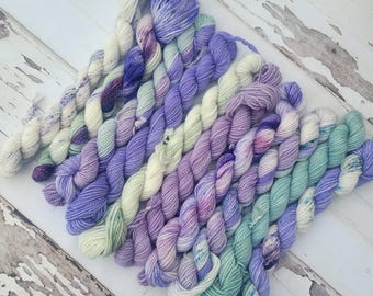 May include: A collection of hand-dyed yarn skeins in various shades of purple, lavender, and teal. The yarn is arranged in a diagonal pattern, showcasing the different color variations and textures. The skeins are displayed on a distressed white wooden surface.