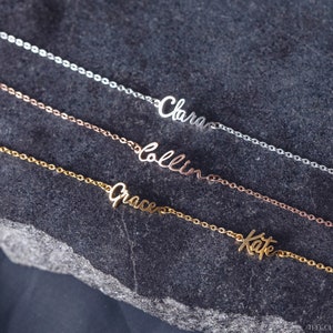 K&ouml;nnte beinhalten: Vier zarte Halsketten aus Gold, Silber und Ros&eacute;gold mit personalisierten Namen, "Clara", "Collin", "Grace" und "Kate", auf einem grauen Steinuntergrund.